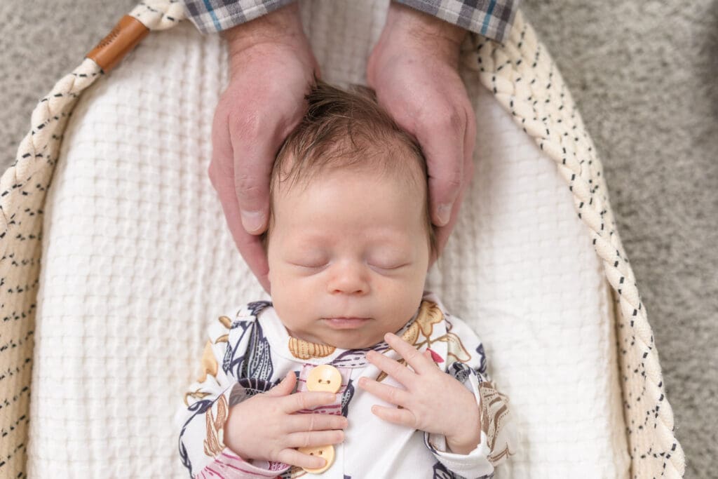 Newborn resting in woven basket with parent hands around baby’s head