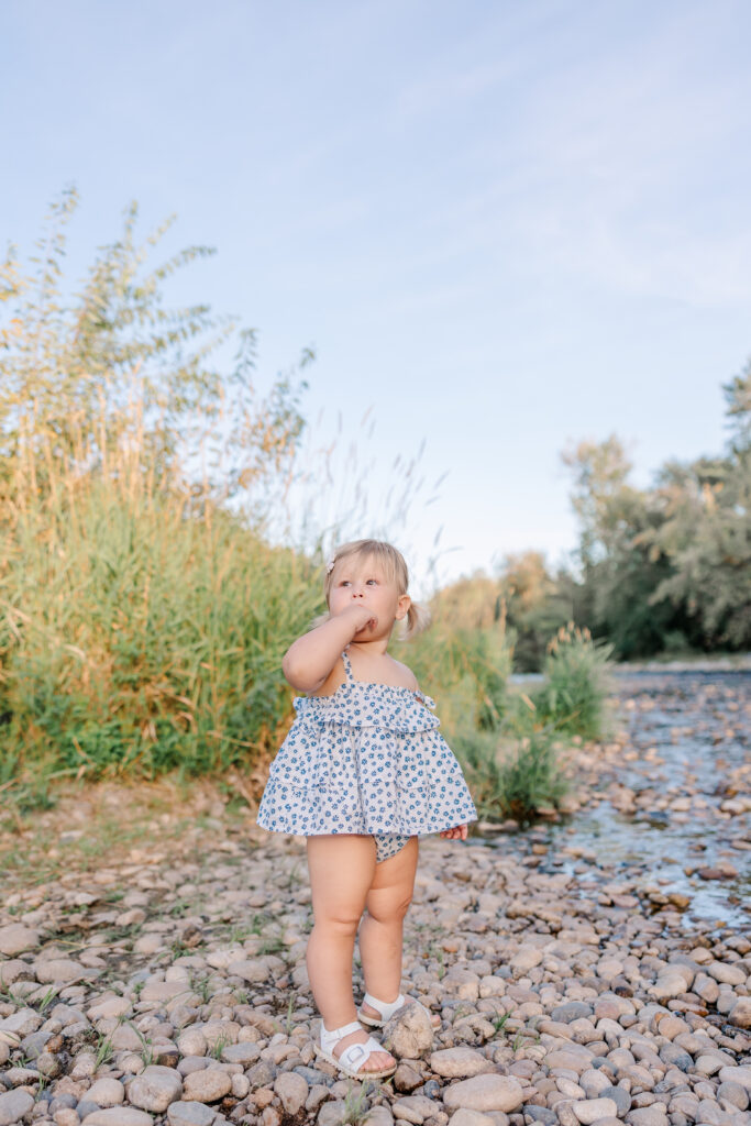 Toddler girl in floral romper standing on rocky riverbank in soft spring light