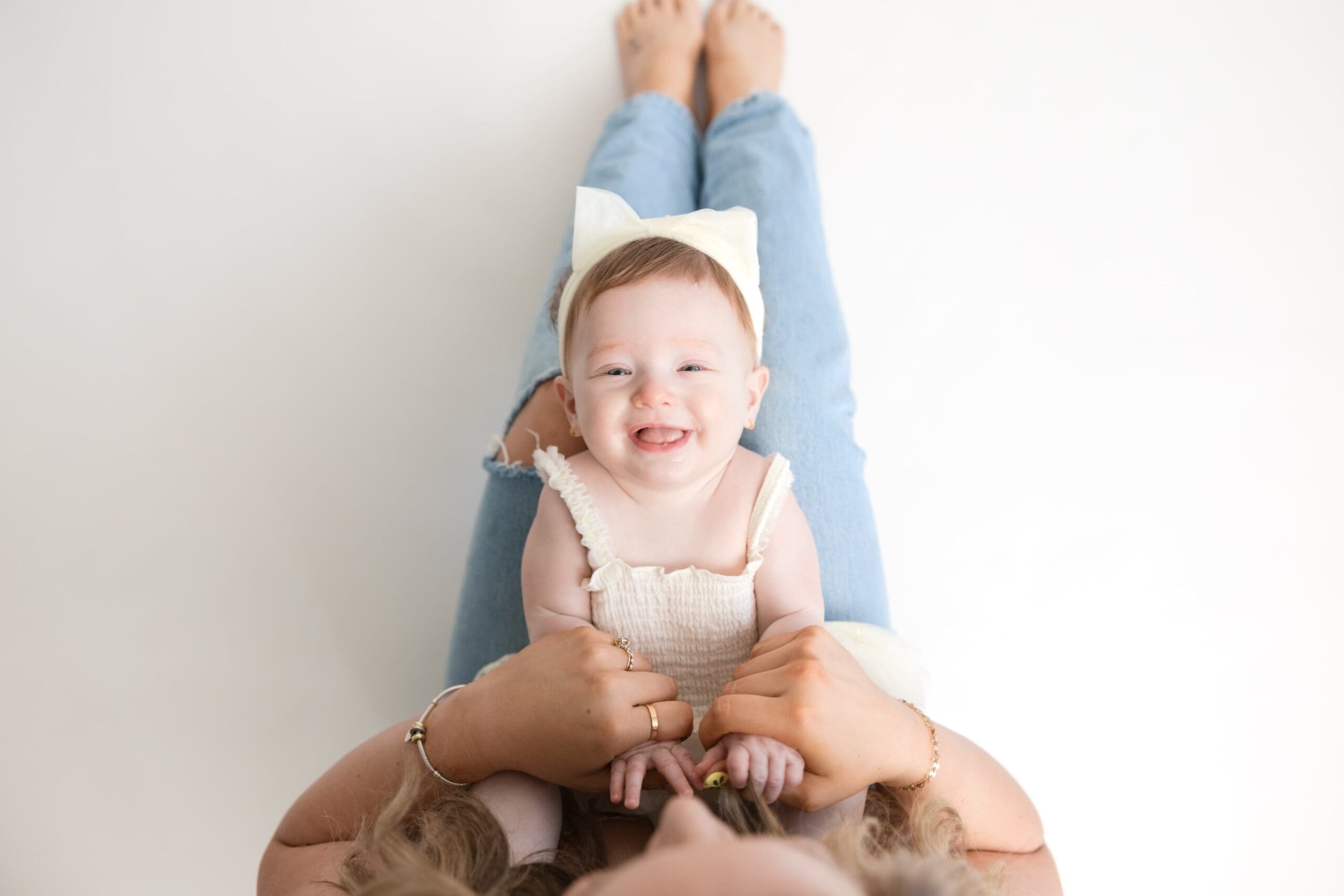 Smiling baby girl in cream outfit lying on her mom’s legs during studio family session