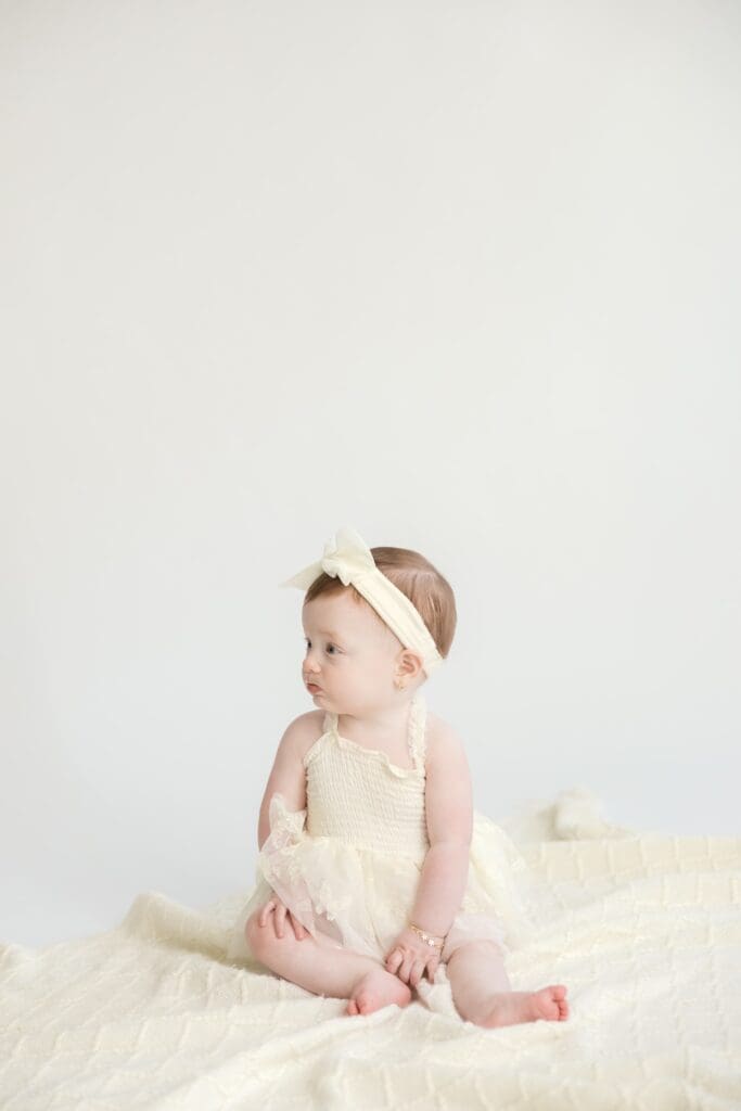 Baby girl in cream dress sitting on soft blanket during milestone session