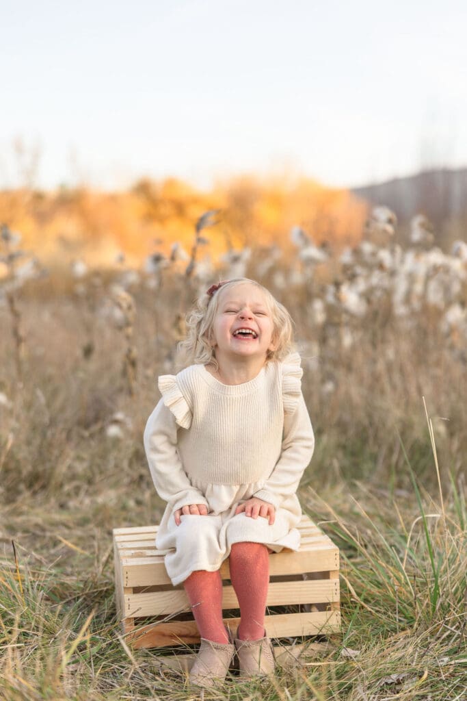 Smiling little girl sitting on a crate surrounded by tall grass during a family photo session